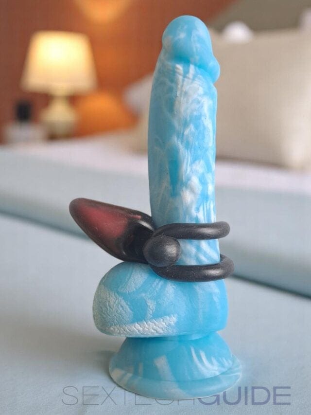Blue and black artistic dildo with abstract design, silicone material, and textured surface for adult pleasure, displayed on a white surface with a blurred bedroom background, product focus for sex toy enthusiasts.