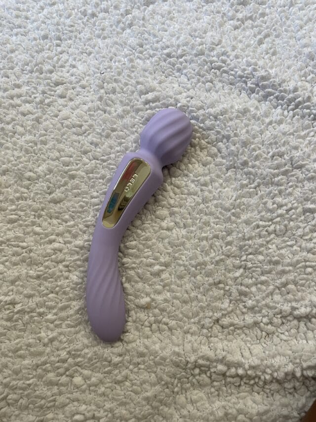 Flexible, lavender-colored sex toy with gold accents lying on a textured beige surface.
