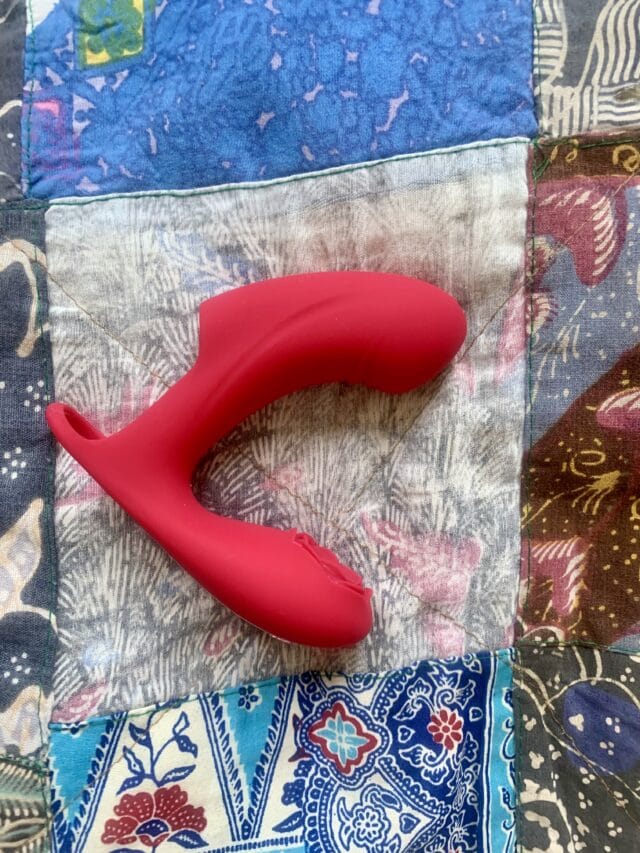 Pink silicone sex toy with a curved design and textured stimulation surface, lying on a colorful patchwork quilt.