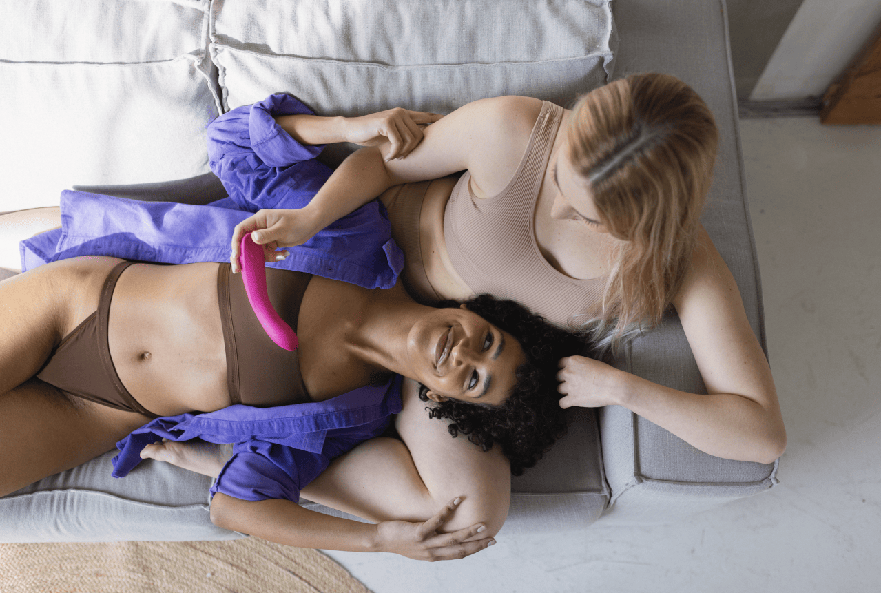 Two women exploring the new We-Vibe Rave 2 dual stimulation vibrator with a twist hinge design while lounging on a couch.